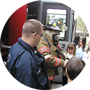 Firefighter in full gear standing by a fire truck talking to a group of small children